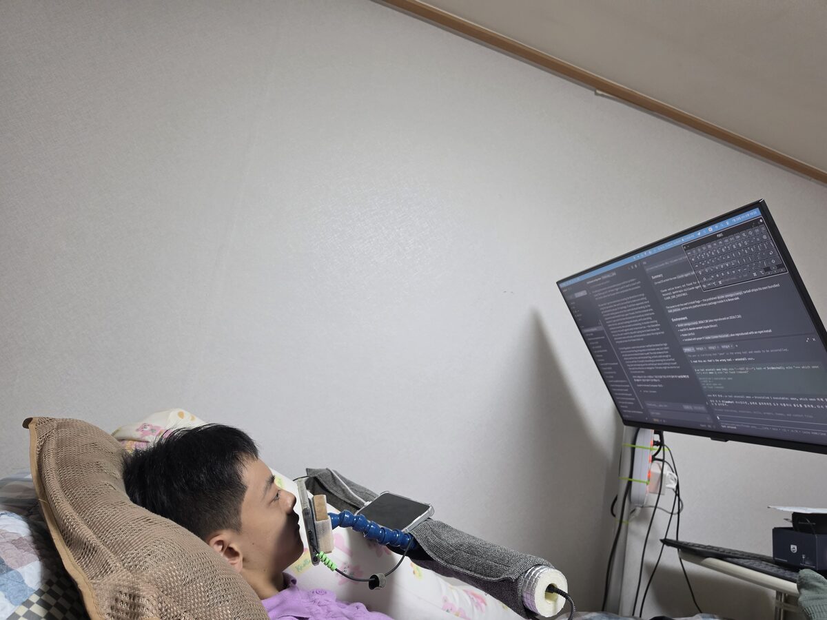 침대에 누워 거치대의 스마트폰 화면을 입술로 눌러 컴퓨터를 조작하는 작업 환경